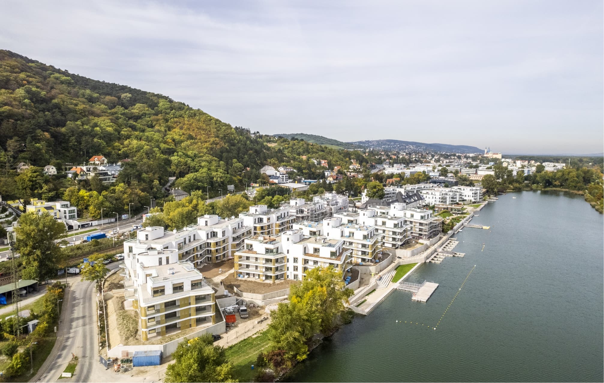 Luftaufnahme eines Wohnensembles am Wasser vor bewaldeten Hügeln